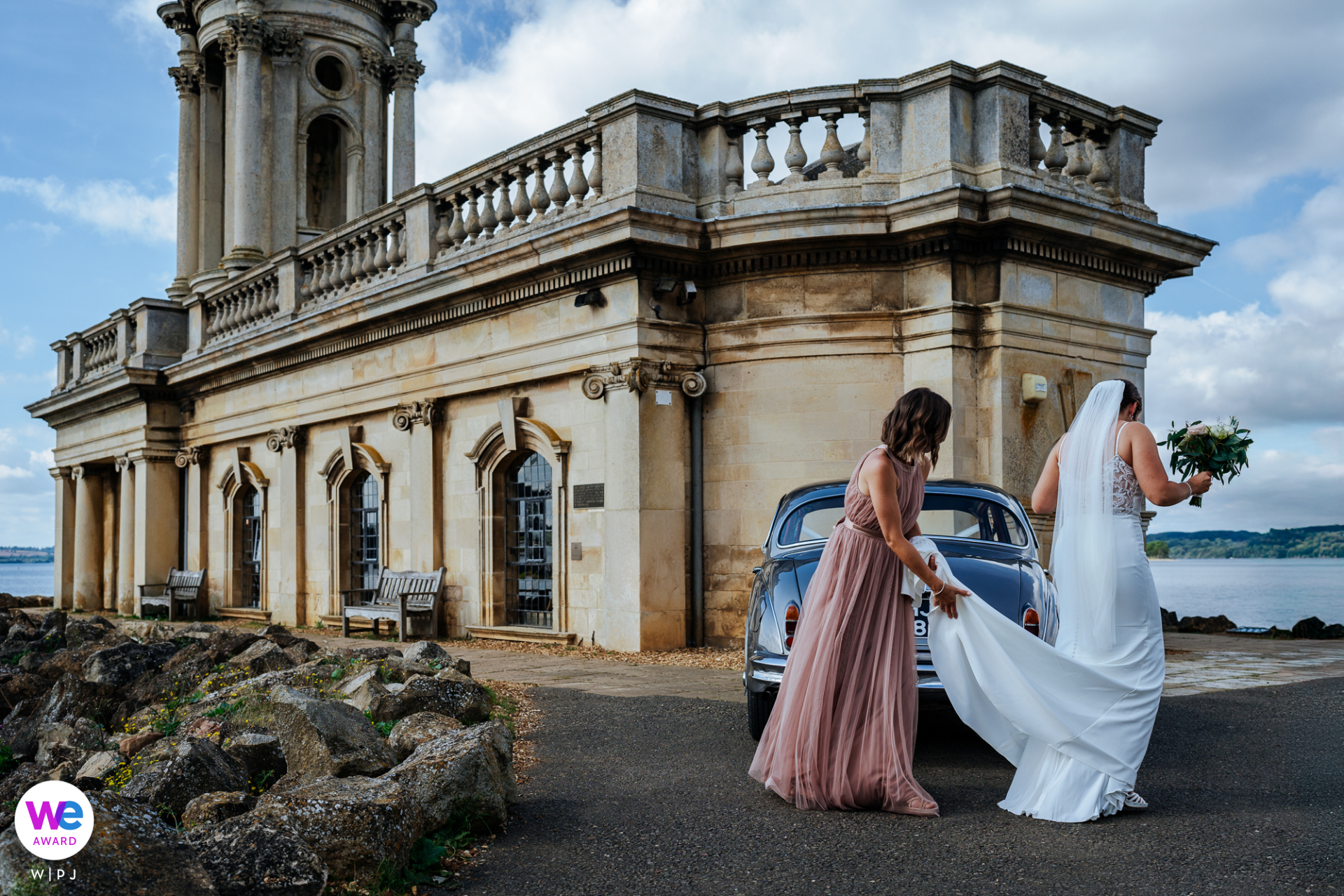 Bride and bridesmaid walking toward an 18th-century church by the scenic water’s edge.